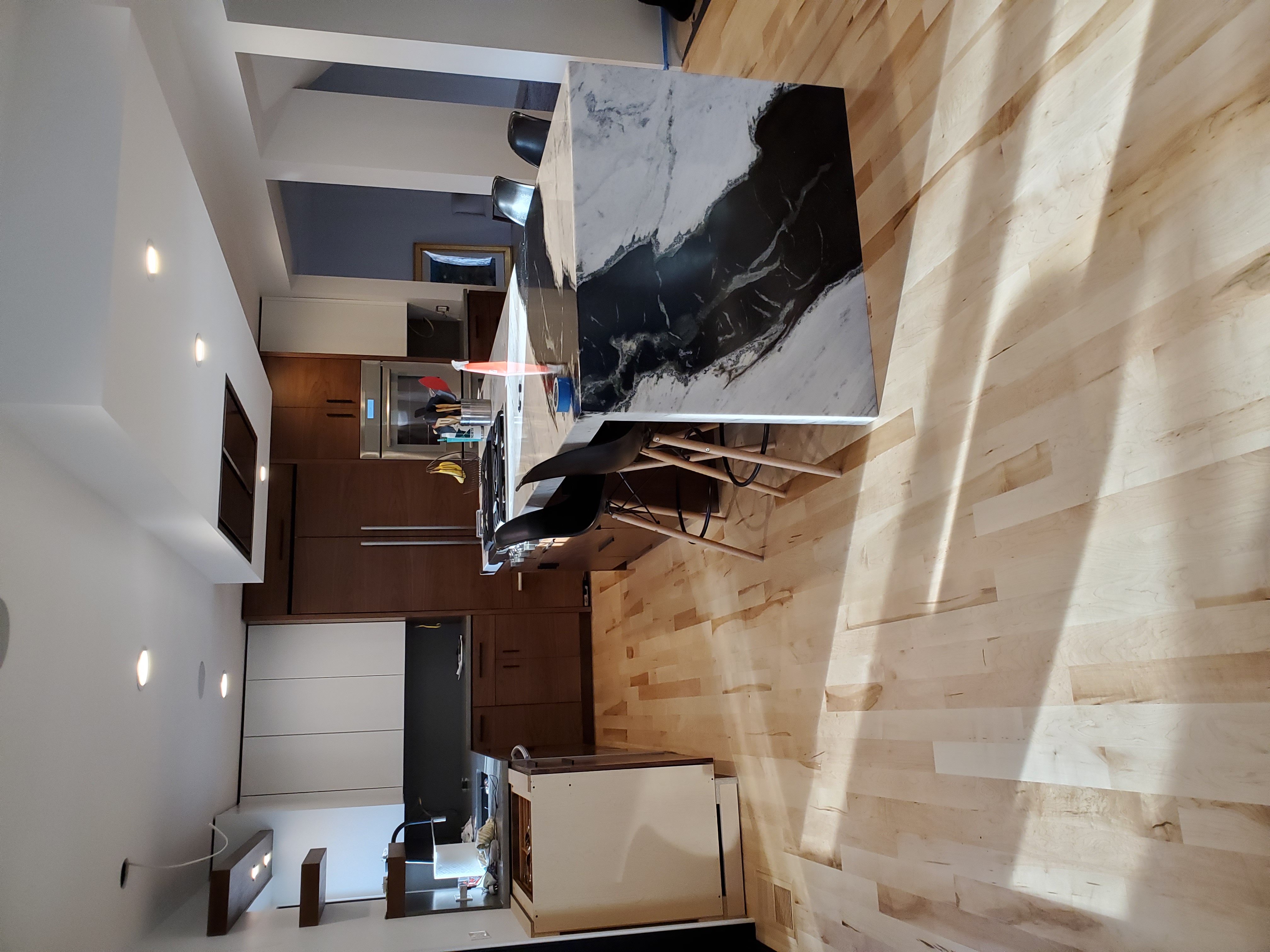 Wide view of the remodeled kitchen with walnut cabinetry, waterfall stone island, and light maple flooring.